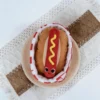 Top view of the Hot Dog Snuffle Plush Dog Toy in a red checkered basket on a plate, showing the soft plush hot dog design for treat hiding and play.
