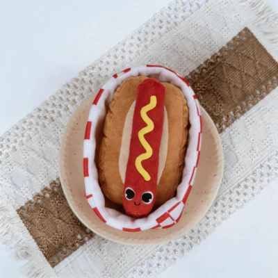Hot Dog Snuffle Plush Dog Toy in Red Checkered Basket Top View Top view of the Hot Dog Snuffle Plush Dog Toy in a red checkered basket on a plate, showing the soft plush hot dog design for treat hiding and play.