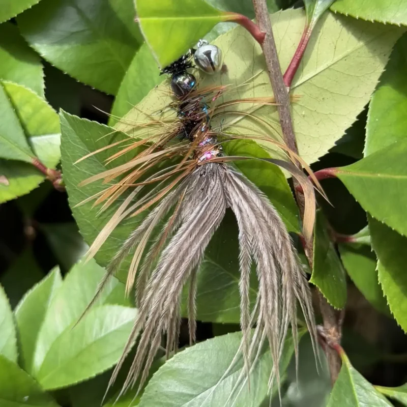 Bird-inspired ostrich feather cat toy resting among green leaves, with soft gray plumes and brown chicken feathers forming a small feathered prey shape.