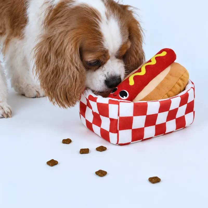 A brown-and-white dog sniffs kibble scattered around the hot dog toy and checkered box during a simple foraging game.