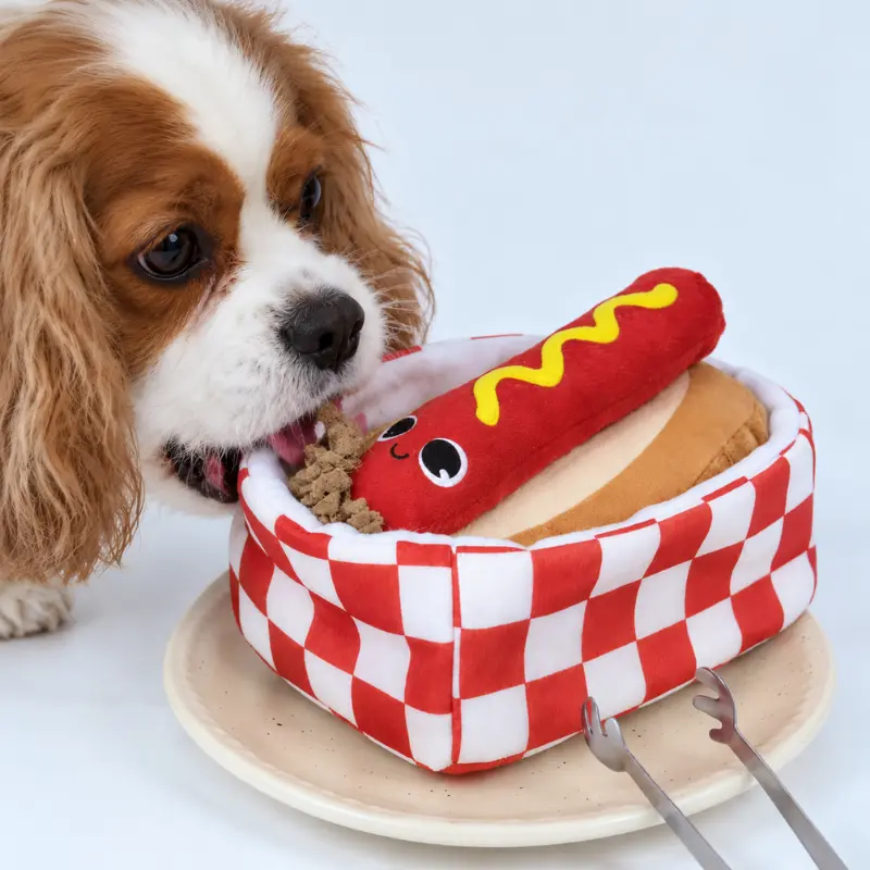 A brown-and-white dog searches for treats in the red-and-white checkered box beside a hot dog toy during interactive treat-finding play.