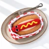 Top-down view of a hotdog plush nestled inside a red-and-white checkered takeaway box on a plate beside a fork.