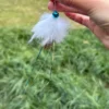 rabbit-fur-cat-toy-iridescent-streamers Hand holding a rabbit fur cat toy attachment with a blue bell and long iridescent streamers against a softly blurred green outdoor background.