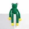 Green floppy dog toy shaped like a frog, sitting on a white display block with wide eyes, yellow paws, and long arms and legs hanging down.