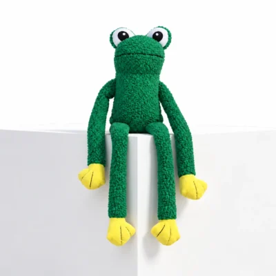 Green floppy dog toy shaped like a frog, sitting on a white display block with wide eyes, yellow paws, and long arms and legs hanging down.