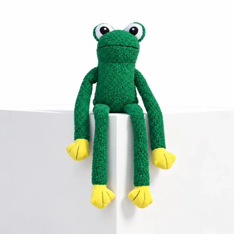 Green floppy dog toy shaped like a frog, sitting on a white display block with wide eyes, yellow paws, and long arms and legs hanging down.