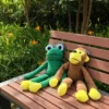 Two dog stuffed animals, a brown monkey and a green frog, sitting together on a wooden garden bench with long floppy limbs and bright yellow paws.