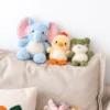 Three Sunfluff stuffed animal dog toys sit together on a sofa: a blue elephant, cream-yellow duck, and sage-green frog, styled as a cozy snuggle stuffed animal trio.