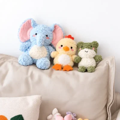 Three Sunfluff stuffed animal dog toys sit together on a sofa: a blue elephant, cream-yellow duck, and sage-green frog, styled as a cozy snuggle stuffed animal trio.