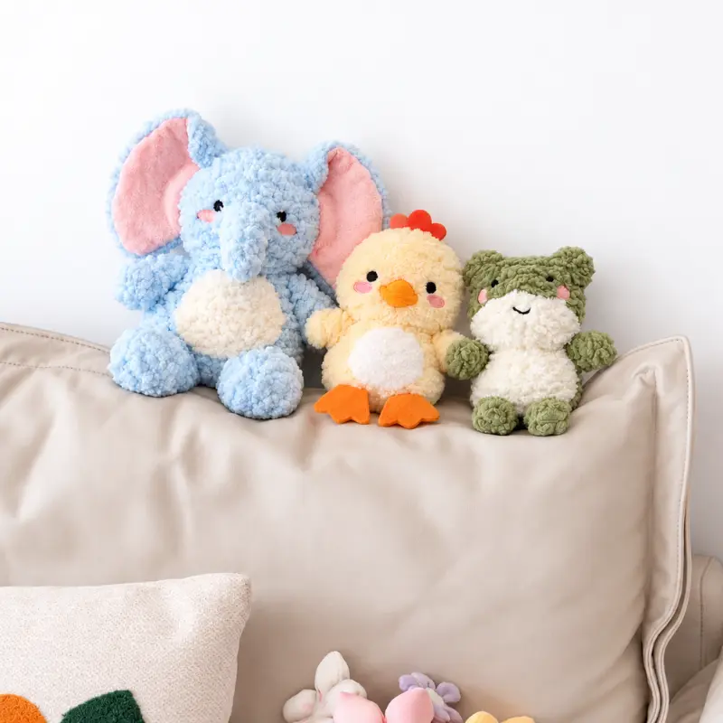 Three Sunfluff stuffed animal dog toys sit together on a sofa: a blue elephant, cream-yellow duck, and sage-green frog, styled as a cozy snuggle stuffed animal trio.